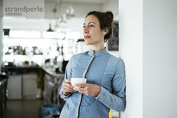 Nachdenkliche Geschäftsfrau mit Kaffeetasse an der Wand eines Cafés
