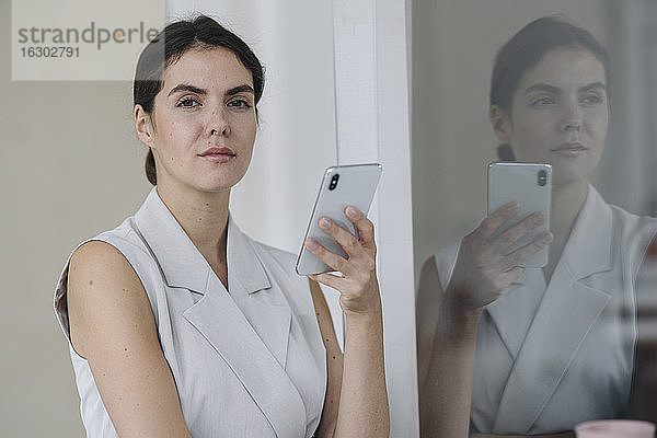 Selbstbewusste Geschäftsfrau  die ein Mobiltelefon benutzt  während sie an einer Glaswand im Büro steht