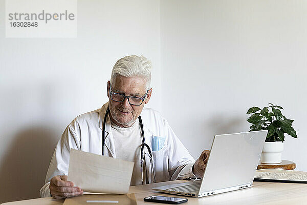 Lächelnder Arzt  der seine Krankenakte betrachtet  während er an der Wand sitzt