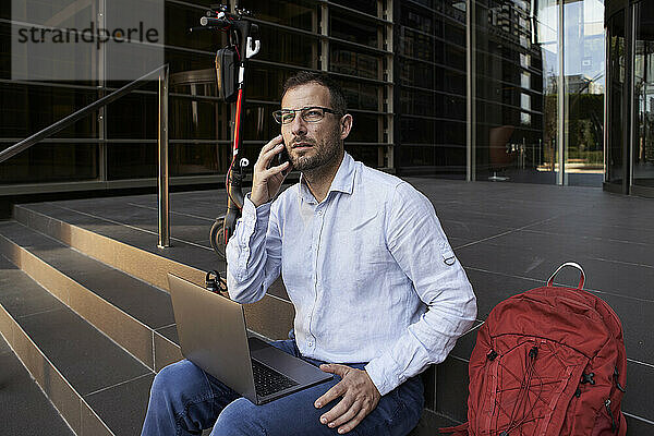 Geschäftsmann mit Laptop  der auf einem Smartphone spricht  während er auf einer Treppe in der Stadt sitzt