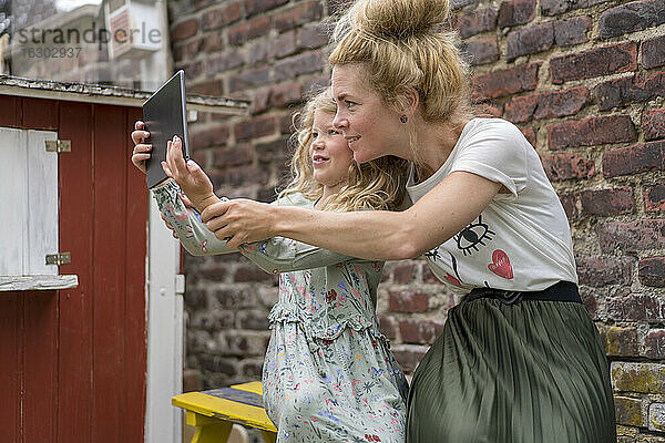 Lächelndes Mädchen nimmt Selfie mit Mutter durch digitale Tablette gegen Backsteinmauer im Hinterhof