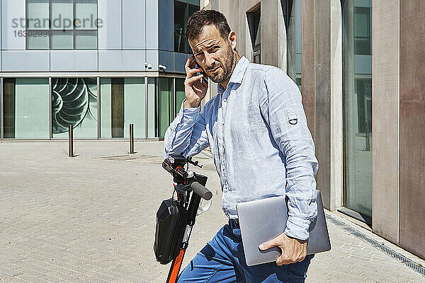 Geschäftsmann telefoniert im Stehen mit Laptop auf einem Motorroller in der Stadt