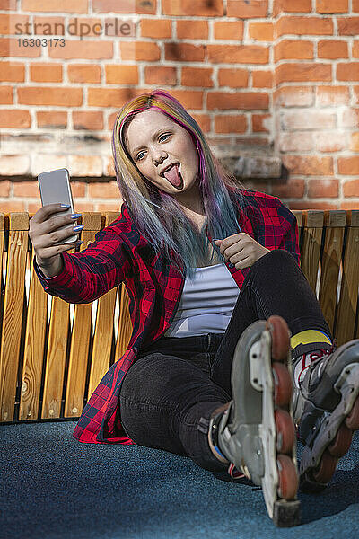 Junge Frau mit gefärbten Haaren und Rollschuhen macht ein Selfie und streckt die Zunge heraus