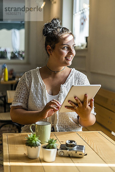 Lächelnde üppige Frau  die ein digitales Tablet benutzt und wegschaut  während sie an einem Tisch im Café sitzt
