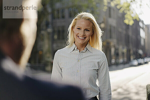Lächelnde blonde Geschäftsfrau  die einen männlichen Kollegen ansieht  während sie in der Stadt steht