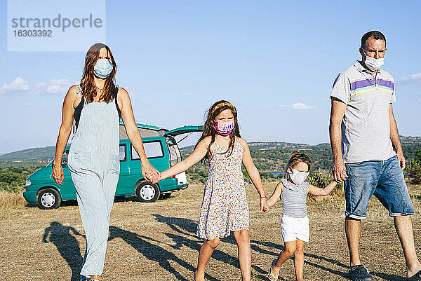 Familie mit Masken  die sich an den Händen halten  während sie durch eine Landschaft gegen den Himmel spazieren