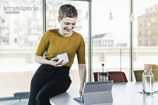 Lächelnde Geschäftsfrau sitzt auf einem Schreibtisch im Büro  hält einen Kaffeebecher und benutzt ein Tablet