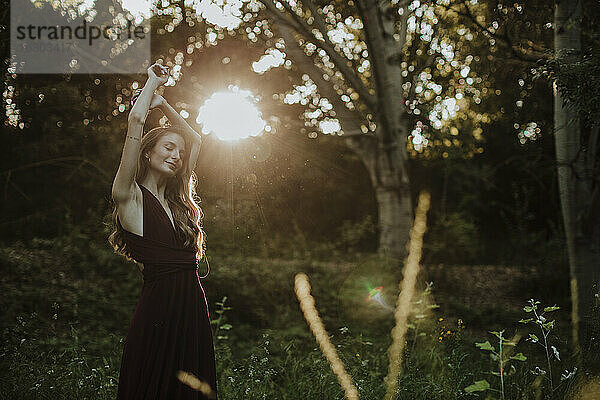 Lächelnde Frau mit gegen das Sonnenlicht erhobenen Armen im Wald stehend
