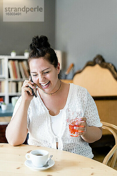 Fröhliche junge Frau  die ein Getränk in der Hand hält und über ein Smartphone spricht  während sie in einem Café sitzt
