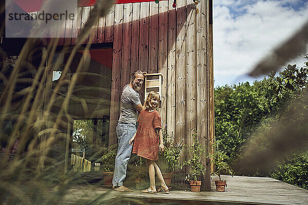 Lächelnder Vater und Tochter schauen weg  während sie ein Insektenhotel an der Wand installieren