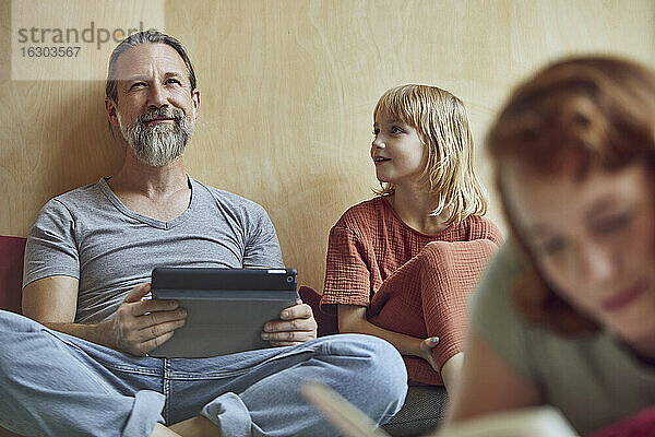 Vater und Tochter mit digitalem Tablet  die wegschauen  während sie zu Hause an der Wand sitzen