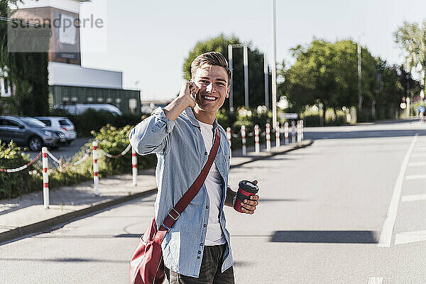 Lächelnder junger Mann  der beim Überqueren einer Straße in der Stadt mit seinem Handy telefoniert