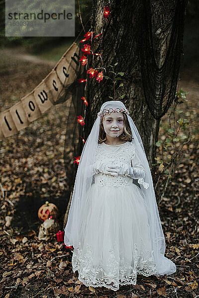 Kleines Mädchen steht im Leichenbrautkostüm vor einem Baum im Wald