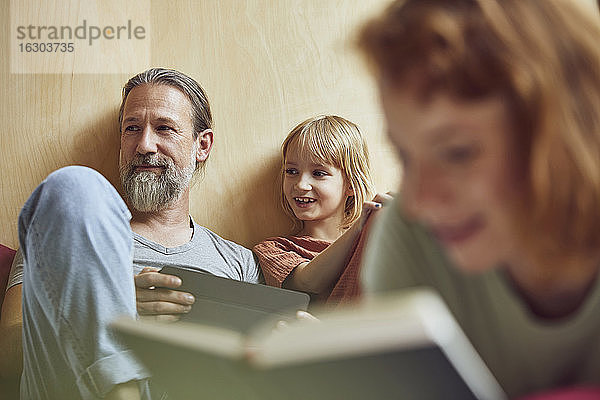 Frau liest ein Buch  während Vater und Tochter zu Hause im Hintergrund wegschauen