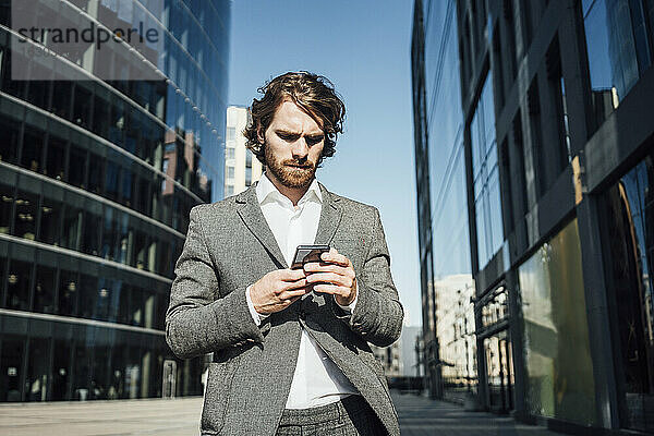 Männlicher Fachmann  der ein Mobiltelefon benutzt  während er an einem modernen Bürogebäude in der Innenstadt an einem sonnigen Tag steht