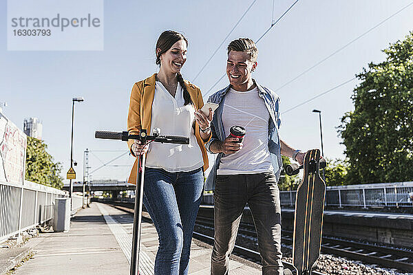 Lächelnde Frau  die einem männlichen Freund beim Spaziergang am Bahnhof ihr Smartphone zeigt