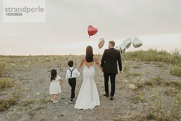 Eltern und Kinder tragen Hochzeitskleid  während sie mit herzförmigen Luftballons im Feld gegen den Himmel laufen
