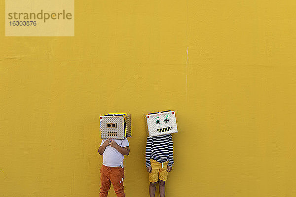 Jungen tragen Robotermasken aus Kartons und stehen an einer gelben Wand