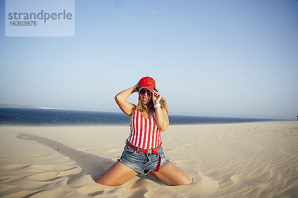 Fröhliche Frau  die eine Mütze hält  während sie am Strand im Sand kniet  gegen einen klaren blauen Himmel