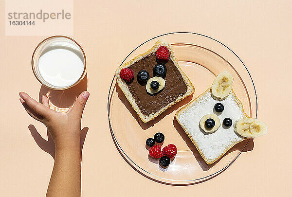 Studioaufnahme von Toasts mit Bärengesichtern aus Früchten und der Hand eines kleinen Mädchens  das nach einem Glas Milch greift