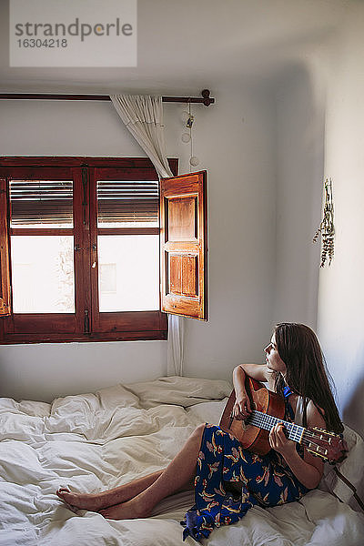 Frau blickt durch das Fenster  während sie im Schlafzimmer zu Hause Gitarre übt