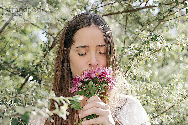 Frau riecht an frischen rosa Blumen  während sie im Frühling in einem Park unter Bäumen steht