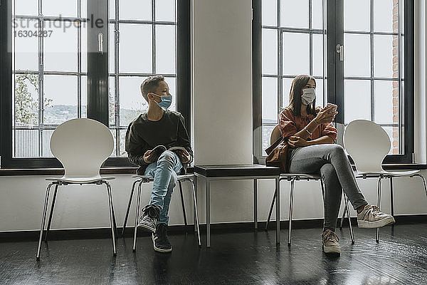 Teenager-Mädchen und Junge mit Masken  die auf Stühlen im Wartezimmer sitzen