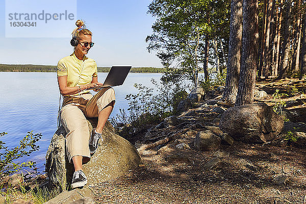 Mittlere erwachsene Frau mit Sonnenbrille  die einen Laptop benutzt  während sie an einem See im Tiveden National Park  Schweden  sitzt