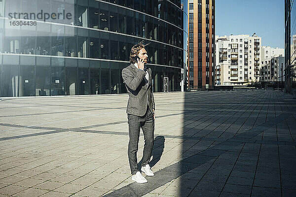 Männlicher Unternehmer  der mit seinem Smartphone telefoniert  während er an einem sonnigen Tag über das Finanzviertel hinwegschaut
