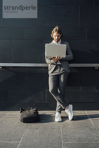 Männlicher Unternehmer  der einen Laptop benutzt  während er an einem sonnigen Tag mit gekreuzten Beinen an der Wand steht