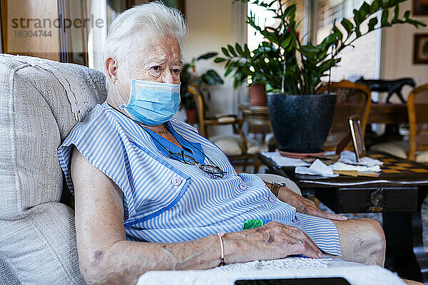 Weibliche Seniorin mit Schutzmaske sitzt zu Hause im Sessel
