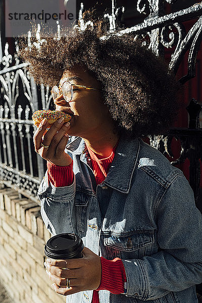 Nahaufnahme einer jungen Frau mit Afro-Haar  die an einem Zaun in der Stadt einen Donut isst