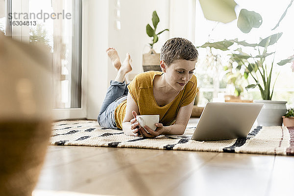 Mittlere erwachsene Frau  die eine Kaffeetasse hält und einen Laptop benutzt  während sie zu Hause auf dem Teppich liegt
