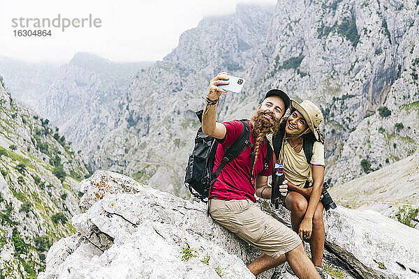 Lächelndes Paar  das ein Selfie macht  während es auf einem Felsen an der Ruta Del Cares sitzt  Asturien  Spanien