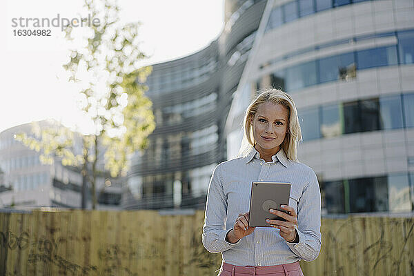 Selbstbewusste Geschäftsfrau  die ein digitales Tablet hält  während sie an einem Gebäude im Finanzviertel steht