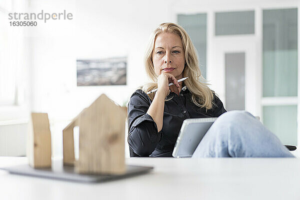 Nachdenkliche Geschäftsfrau mit digitalem Tablet  die ein Modell auf dem Schreibtisch betrachtet  während sie in ihrem Büro zu Hause sitzt