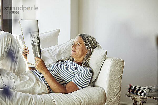 Frau liest eine Zeitschrift  während sie auf dem Sofa im Wohnzimmer liegt