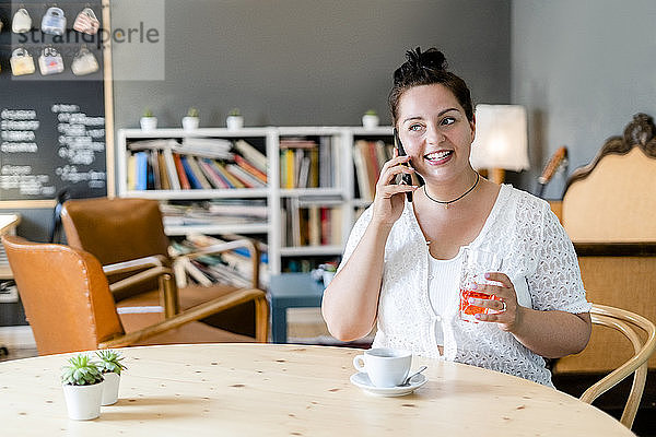 Üppige Frau  die ein Getränk hält und über ein Smartphone spricht  während sie in einem Café sitzt