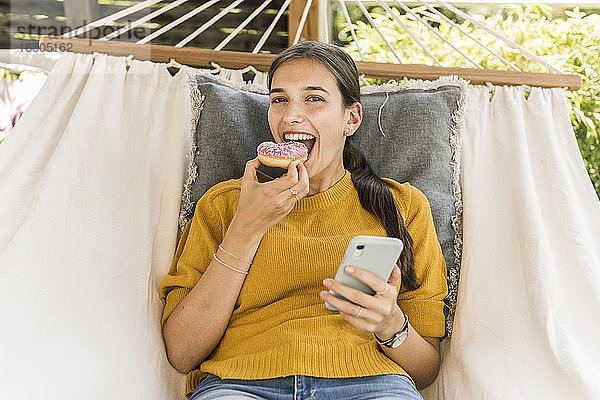 Fröhliche junge Frau isst Donut  während sie in der Hängematte ihr Handy benutzt