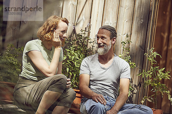 Lächelndes Paar  das sich unterhält  während es bei Topfpflanzen vor einem kleinen Haus sitzt
