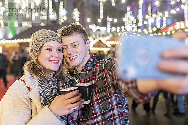 Paar hält heiße Schokolade und macht ein Selfie auf dem nächtlichen Weihnachtsmarkt