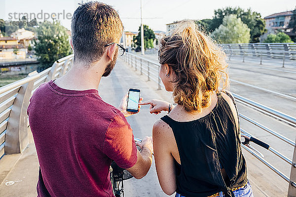 Ein Paar benutzt ein Smartphone  während es mit Fahrrädern auf einer Brücke bei Sonnenuntergang steht