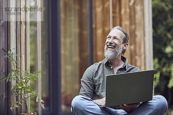 Fröhlicher reifer Mann mit Laptop  der wegschaut  während er vor einem kleinen Haus sitzt