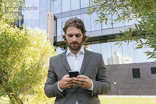Hübscher Geschäftsmann mit Bart  der ein Smartphone benutzt  während er vor einem Bürogebäude steht