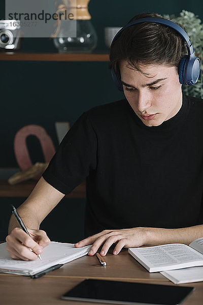 Hübscher junger Student  der in ein Buch schreibt  während er zu Hause über Kopfhörer Musik hört