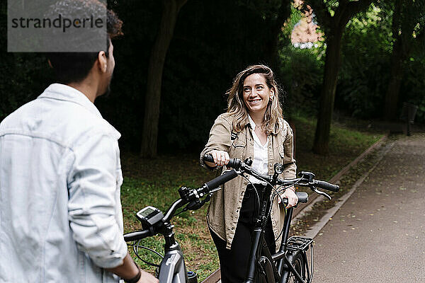 Lächelnde junge Frau sieht ihren Freund an  während sie mit einem Elektrofahrrad auf der Straße steht