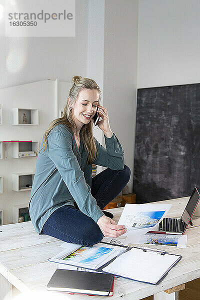 Lächelnde Frau sitzt auf dem Schreibtisch im Home Office und telefoniert
