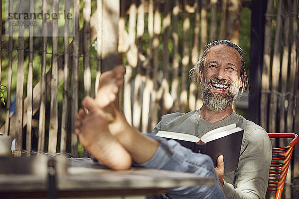 Fröhlicher bärtiger Mann liest ein Buch und entspannt sich auf einem Stuhl im Garten