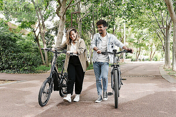 Lächelnder Freund und Freundin gehen mit Elektrofahrrädern auf der Straße