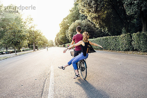 Junge Frau mit ausgestreckten Armen sitzt mit ihrem Freund auf einem Fahrrad im Park